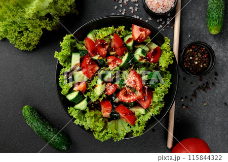 Vegetable salad of tomatoes, cucumbers, onions, lettuce dressed with aromatic dressing in a black plate on a gray background 115844322