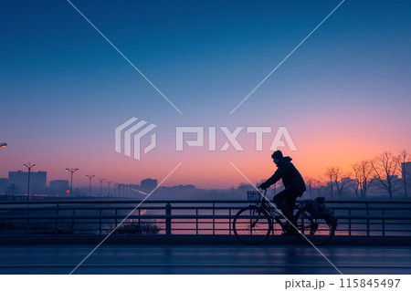 Man biking across bridge at sunrise with city skyline in background 115845497