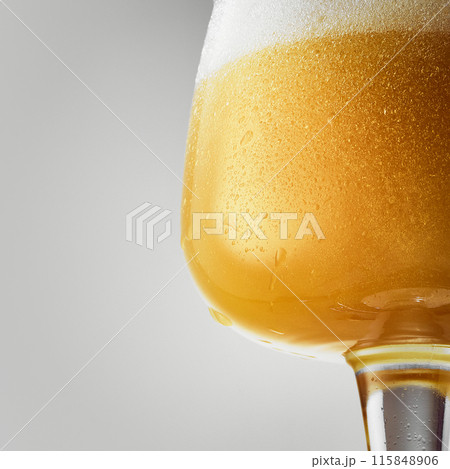 Close-up of mug filled with golden, hazy beer topped with thick, frothy white foam against grey studio background. Close-up of mug filled with golden, hazy beer topped with thick, frothy white foam against grey studio background. 115848906