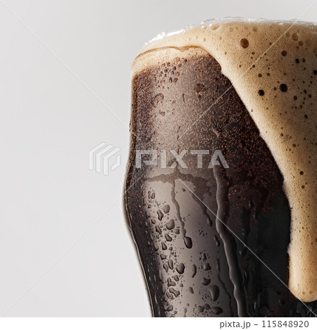 Close-up of pint glass of dark beer, with a creamy, overflowing head of foam against grey studio background. 115848920