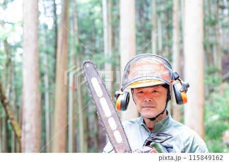 手入れをした森をバックにチェンソーを携えて正面を見るフォレストワーカー 115849162
