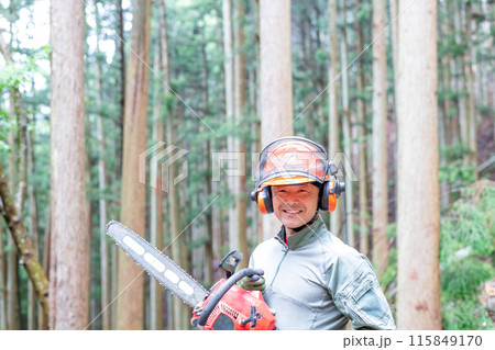 手入れをした森をバックにチェンソーを携えながら笑顔で正面を向くフォレストワーカー 115849170