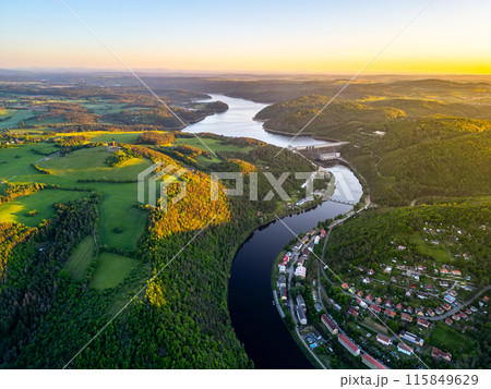 A sunset aerial shot of Orlik Reservoir in Czechia shows a winding river, hills, and a quaint town in the valley with vibrant blue and golden skies. A sunset aerial shot of Orlik Reservoir in Czechia shows a winding river, hills, and a quaint town in the valley with vibrant blue and golden skies. 115849629