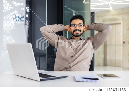 A confident businessman taking a relaxing break while sitting at his desk in a modern office with a laptop, notepad, and tablet. The man appears content and thoughtful in a professional setting. A confident businessman taking a relaxing break while sitting at his desk in a modern office with a laptop, notepad, and tablet. The man appears content and thoughtful in a professional setting. 115851188