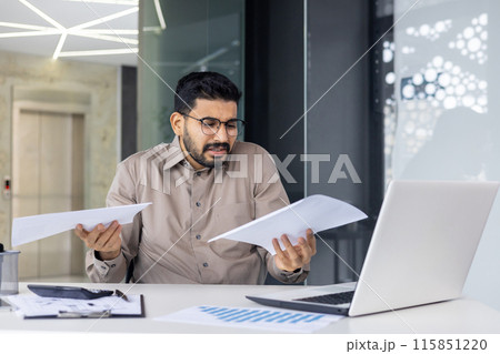 A stressed businessman sits at his desk, looking confused and frustrated with paperwork in his hands. He is working on a laptop in a modern office setting, dealing with financial documents. 115851220