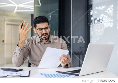 Young man wearing glasses and looking stressed and frustrated while holding paperwork in an office setting. He is seated at a desk with a laptop, documents. Emotion of worry and confusion. 115851225
