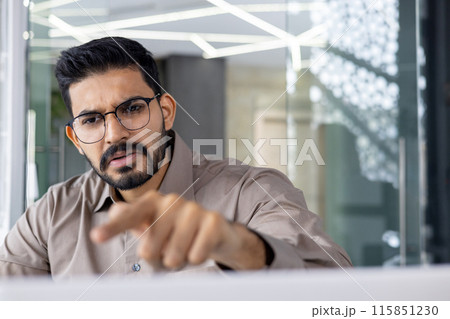 Angry business man with glasses and beard, dressed in a beige shirt, expressing frustration while pointing at his computer screen in a modern office environment. 115851230