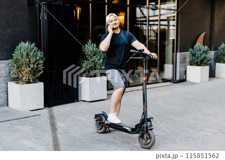 Caucasian young handsome man standing on street at electric scooter and talking on mobile phone. Stylish guy speaking on cellphone outdoors 115851562