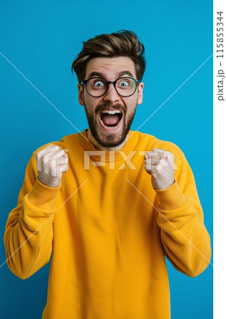 Excited Young Man in Yellow Sweater Celebrating Success on Blue Background 115855344