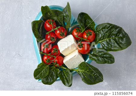 Cherry tomatoes, brine cheese and spinach in a blue decorative bowl on a gray stone table. Top view with a copy of the space 115856074