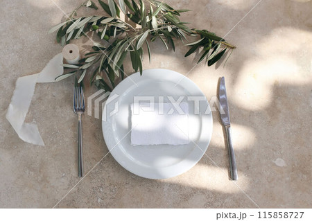 Summer wedding stationery. Blank business card, invitation on white ceramic plate. Olive tree branches, silver fork, knife. Festive table setting. Beige marble background in sunlight. Flat lay, top. 115858727