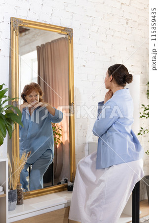 Young attractive woman looks at mirror and see elderly woman, mature herself from future, smiling joyfully to her. Young attractive woman looks at mirror and see elderly woman, mature herself from future, smiling joyfully to her. 115859435