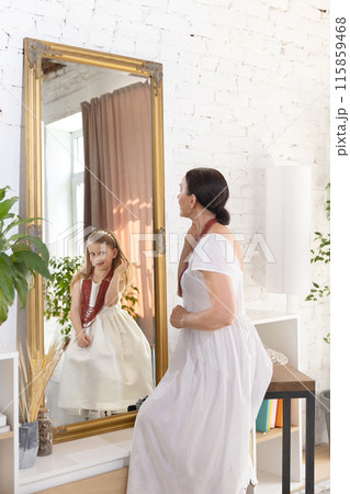 Yung again in another world. Elderly woman in white dress looks at her reflection in mirror, where young girl in matching dress smiles back at her. 115859468