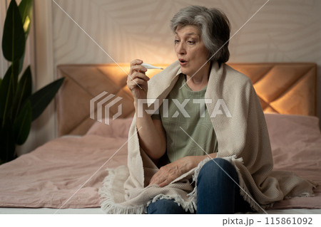 An elderly woman is sitting in bed, wrapped in a blanket, and looks sick. It seems that she measures the temperature of her head with a thermometer. The scene conveys a sense of discomfort and 115861092