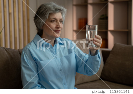 An elderly woman is sitting on the couch in her living room and drinking water from a glass. She enjoys the cool water on a warm day. 115861141
