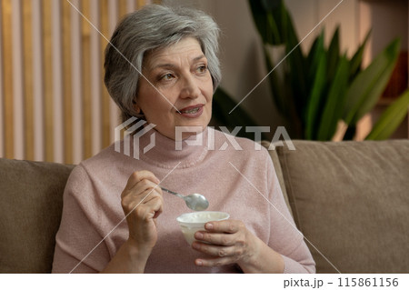 Elderly Woman Eats Yogurt While Sitting On A Couch In The Evening 115861156