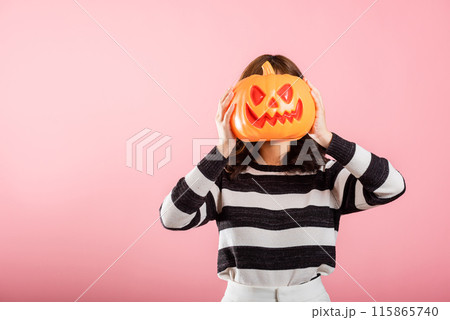 A portrait of a joyful Asian woman holding model pumpkins on her head in a Halloween-themed studio shot. She's all smiles with ghost pumpkins on a pink background. A portrait of a joyful Asian woman holding model pumpkins on her head in a Halloween-themed studio shot. She's all smiles with ghost pumpkins on a pink background. 115865740