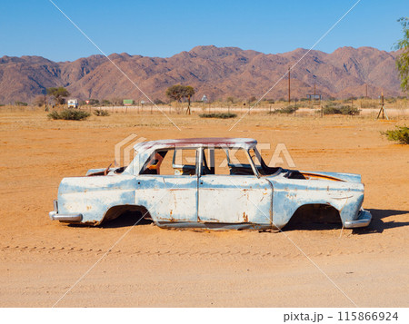 A weathered, rusty car sits abandoned against the arid backdrop of Solitaire, Namibia, under a clear blue sky. 115866924