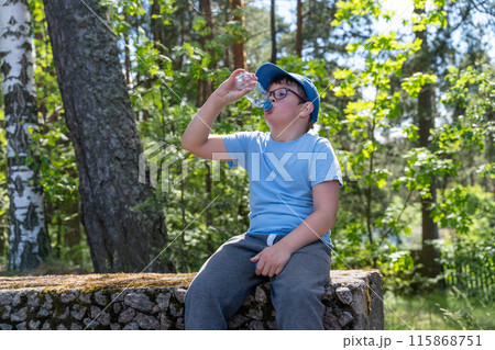 A boy on stone wall in forest drinks water, sun through trees 115868751