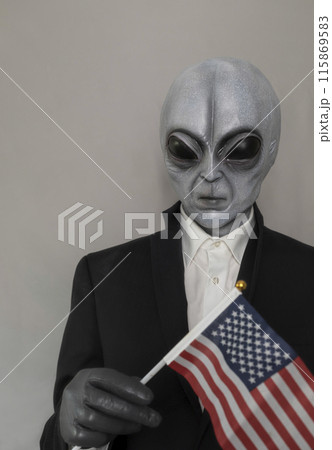Extra Terrestrial Being, Portrait Of Alien Wearing Suit And Holding American Usa Flag on Gray Background. World UFO Day, Unidentified Flying Objects. Vertical Plane. Grey Humanoid. 115869583