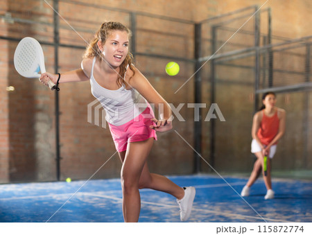 Expressive energetic girl playing padel on indoor court 115872774