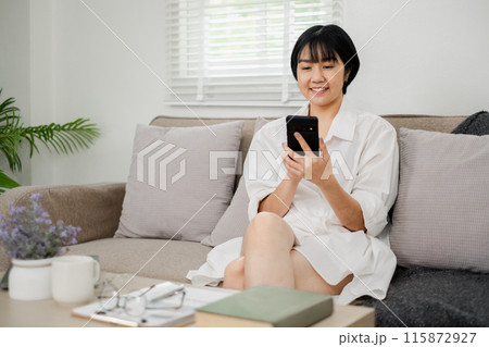 Young woman sitting on a sofa using a smartphone in a bright, modern living room with minimalist decor and natural light. 115872927