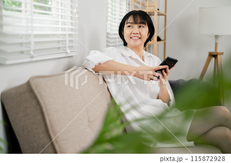 A woman in a white shirt sits on a beige sofa holding a smartphone, smiling and looking away in a bright, modern living room. A woman in a white shirt sits on a beige sofa holding a smartphone, smiling and looking away in a bright, modern living room. 115872928