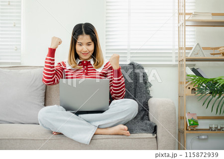 Young woman in casual clothes celebrating success while working on a laptop at home, sitting on a sofa in a bright and modern living room. 115872939