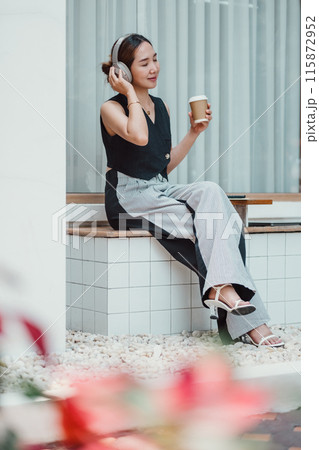 A young woman wearing headphones and holding a coffee cup, sitting in a modern outdoor setting, enjoying music and relaxing. A young woman wearing headphones and holding a coffee cup, sitting in a modern outdoor setting, enjoying music and relaxing. 115872952