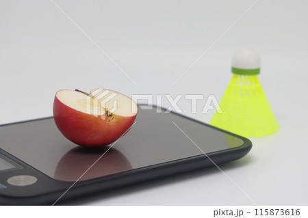 An apple on a kitchen scale against a background of a shuttlecock. Calorie counting An apple on a kitchen scale against a background of a shuttlecock. Calorie counting 115873616