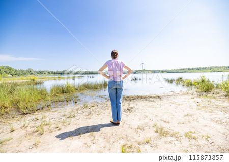 A woman stands by the lakeside, gazing at a serene landscape of a wind turbine in the background 115873857