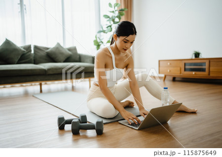 Young Asian woman sitting on the floor on yoga mat in front of laptop and training at home Young Asian woman sitting on the floor on yoga mat in front of laptop and training at home 115875649