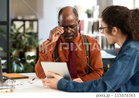 Portrait of African American senior man adjusting glasses while looking at tablet screen in business meeting with young colleague in office copy space Portrait of African American senior man adjusting glasses while looking at tablet screen in business meeting with young colleague in office copy space 115878163
