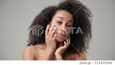 Beauty and healthcare concept - beautiful African American woman with curly afro hairstyle and clean, healthy skin touches her cheek and face with her hand, posing and looking at the camera 115879300
