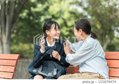 公園のベンチで笑顔で会話する女性・電話で朗報を受ける制服姿・ブレザー姿の女・母娘・中学生・教師・先生 115879474
