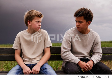 Two teenagers sitting on a bench under a stormy sky, engaging in a serious conversation, with a natural landscape in the background 115879901