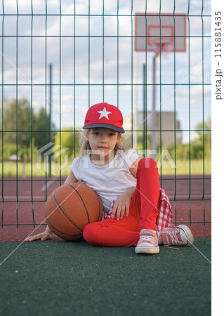 A child in a red baseball cap sits with a basketball on the playground resting after game A child in a red baseball cap sits with a basketball on the playground resting after game 115881435
