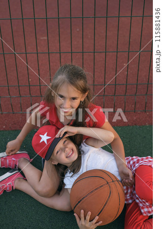 Beautiful preschool girls relax after playing basketball, sitting on the playground with a ball Beautiful preschool girls relax after playing basketball, sitting on the playground with a ball 115881436