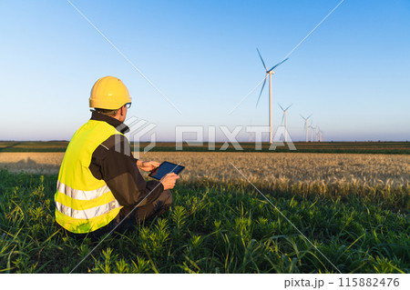 Worker with digital tablet sits on the grass and looks at wind turbines. 115882476