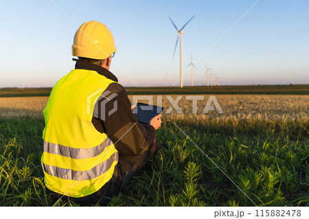 Worker with digital tablet sits on the grass and looks at wind turbines. Worker with digital tablet sits on the grass and looks at wind turbines. 115882478