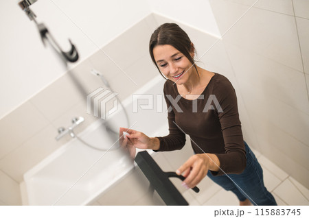 Dark-haired woman cleaning the shower cabin and looking contented Dark-haired woman cleaning the shower cabin and looking contented 115883745