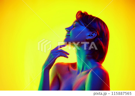 Woman with short brown hair tilts her head back, closing her eyes and gently touching her neck in neon light against gradient studio background. Woman with short brown hair tilts her head back, closing her eyes and gently touching her neck in neon light against gradient studio background. 115887565