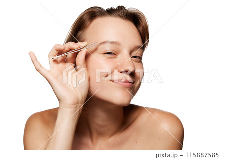 Young woman with short brown hair smiles while holding tweezers near her eyebrow, as if grooming it against white studio background. 115887585