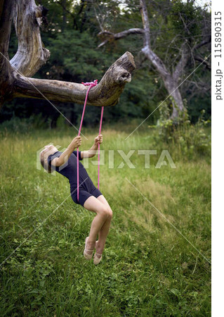 a little 5-year-old gymnast in a black minimalist jumpsuit swings on a gymnastic rope on a tree 115890315