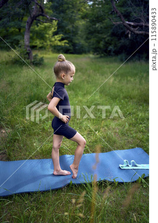 little 5-year-old girl in a black minimalist overall does gymnastics in the garden, outdoors 115890433