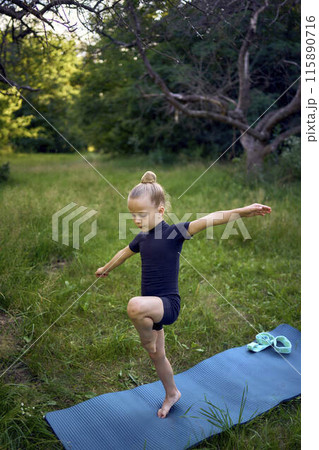 a little 5-year-old girl in a black minimalist jumpsuit does gymnastics in the garden, outdoors 115890716