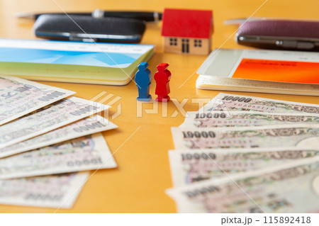 2つの通帳と印鑑、一万円札、家を前に立つ男性と女性のミニチュア、住宅ローン ペアローンのイメージ 115892418