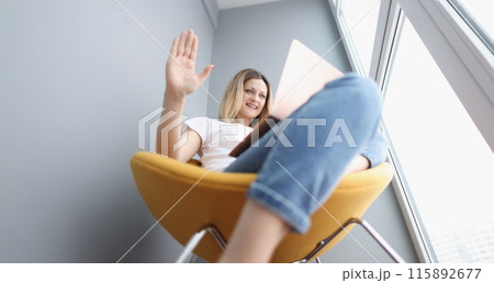 Young woman sitting on armchair and waving hand at laptop screen Young woman sitting on armchair and waving hand at laptop screen 115892677
