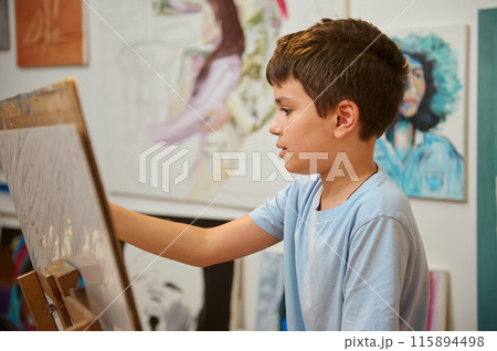 A young boy is diligently working on a painting during an art class. He is focused and engaged in the creative process with other artworks in the background. 115894498