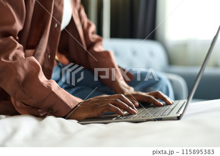 Side view closeup of African American young woman typing on laptop keyboard while sitting on bed in hotel room copy space Side view closeup of African American young woman typing on laptop keyboard while sitting on bed in hotel room copy space 115895383